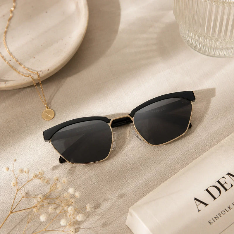 Aesthetic flat lay of LADDER black-and-gold geometric sunglasses on cream linen, styled with a gold necklace, dried florals, and an open book.
